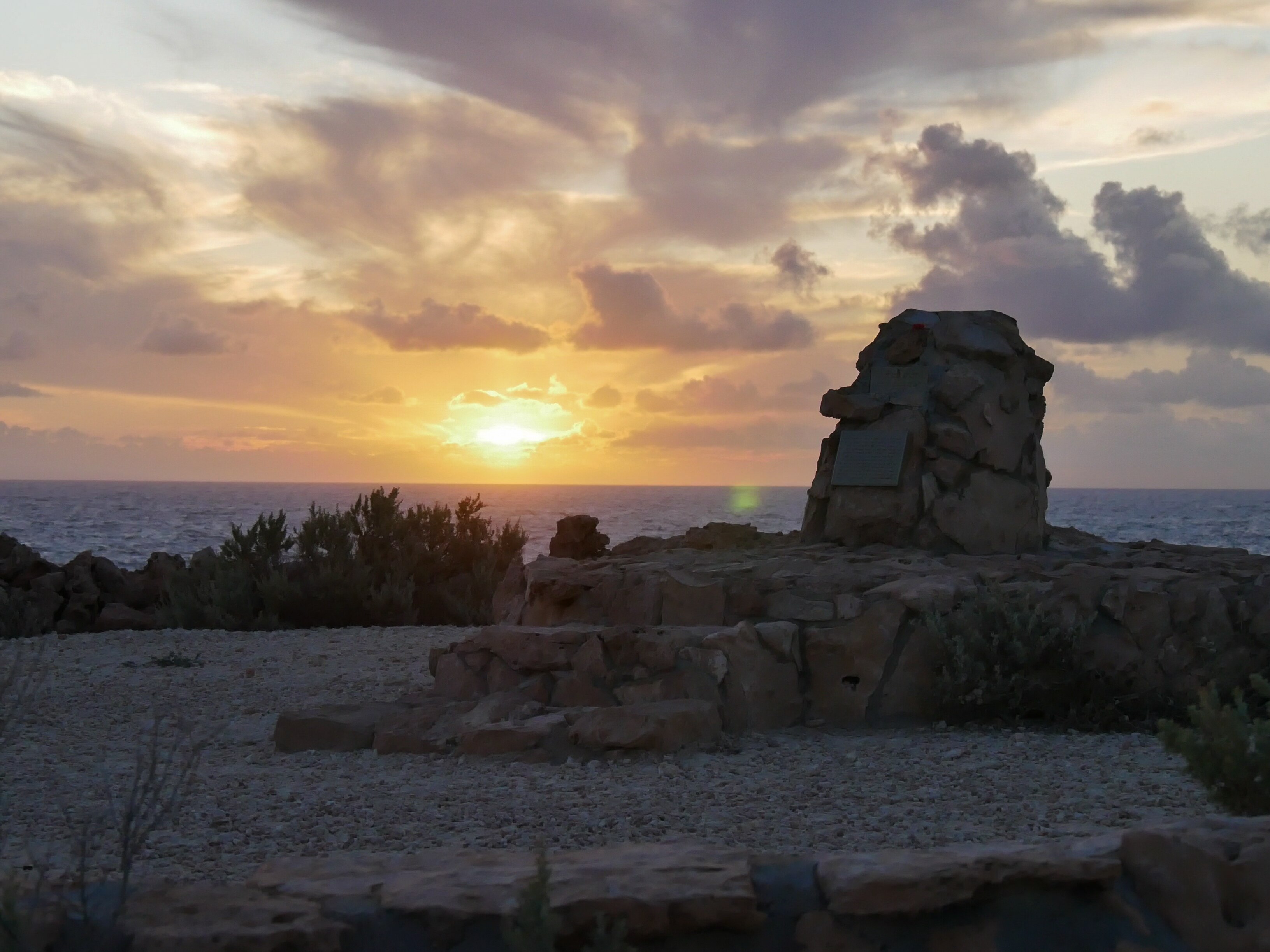 Sun behind clouds above an ocean horizon and a rocky monument in the foreground.