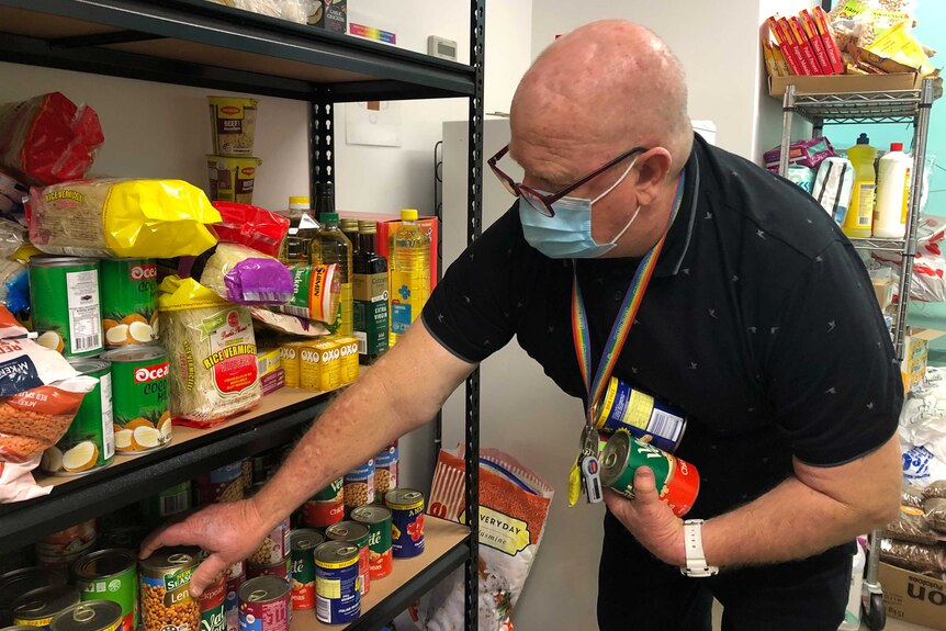 A man wearing a surgical mask puts cans of food and other grocieries on a shelf.