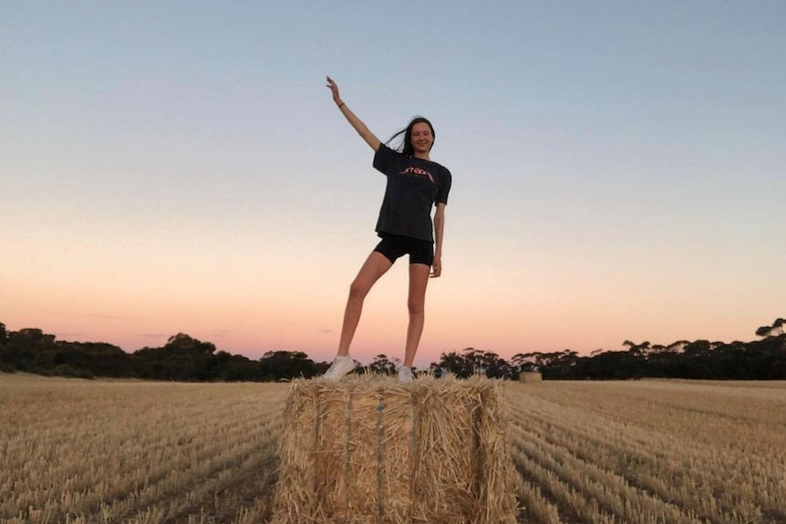 A woman wearing a black T-shirt and black shorts standing on a hay bale among reaped hay