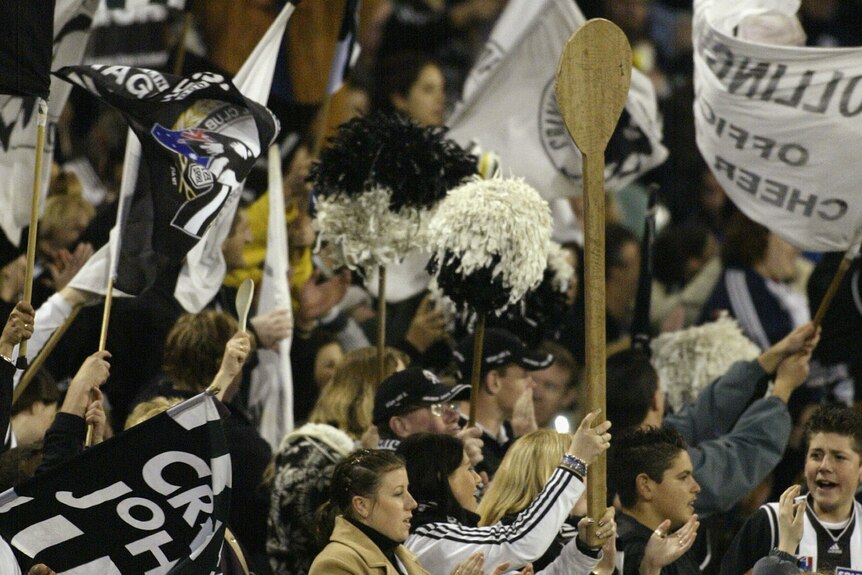 A big group of Collingwood AFL fans celebrate in the stands,  as one woman holds a giant wooden spoon above her.