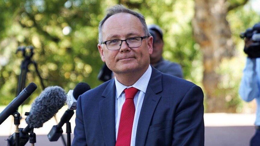 A Minister wearing a suit and tie smiles at the camera with microphones and media in the background.
