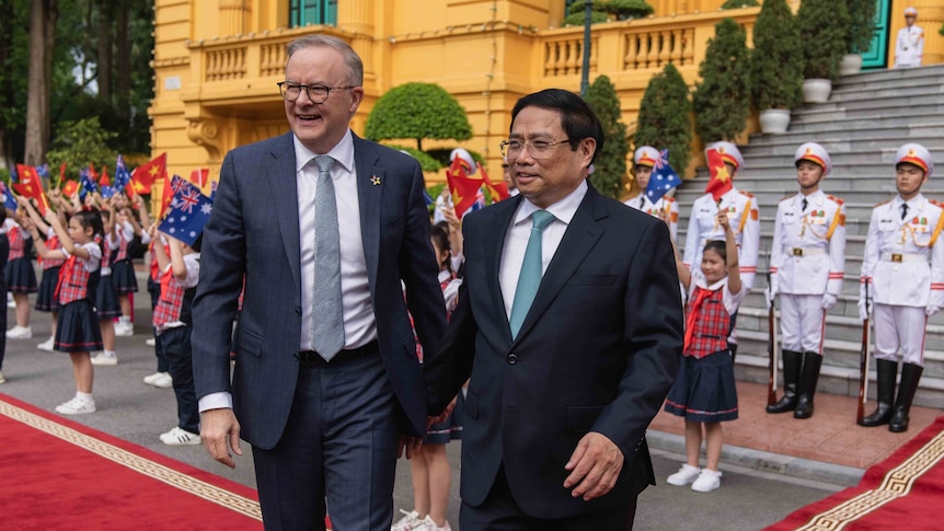Anthony Albanese and Pham Minh Chinh standing in front of a building with military and young Vietnamese children holding flags.