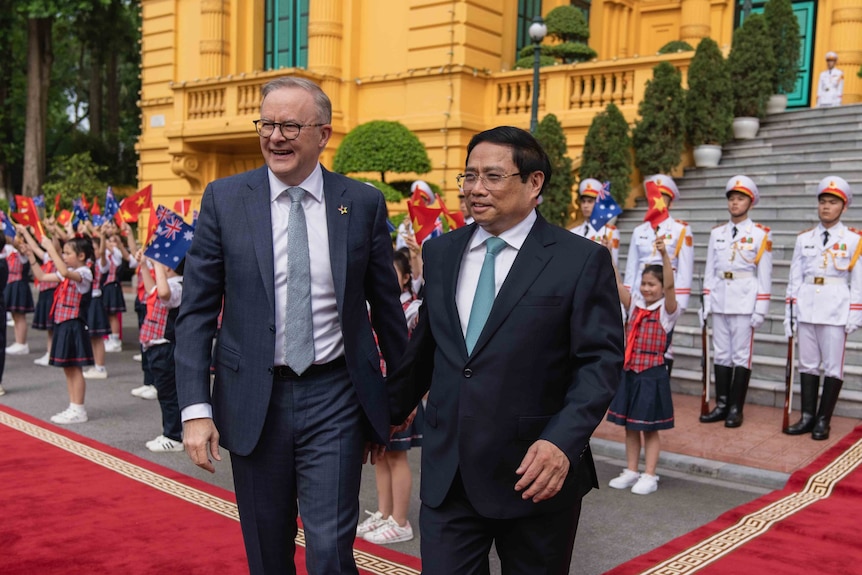 Anthony Albanese and Pham Minh Chinh standing in front of a building with military and young Vietnamese children holding flags.