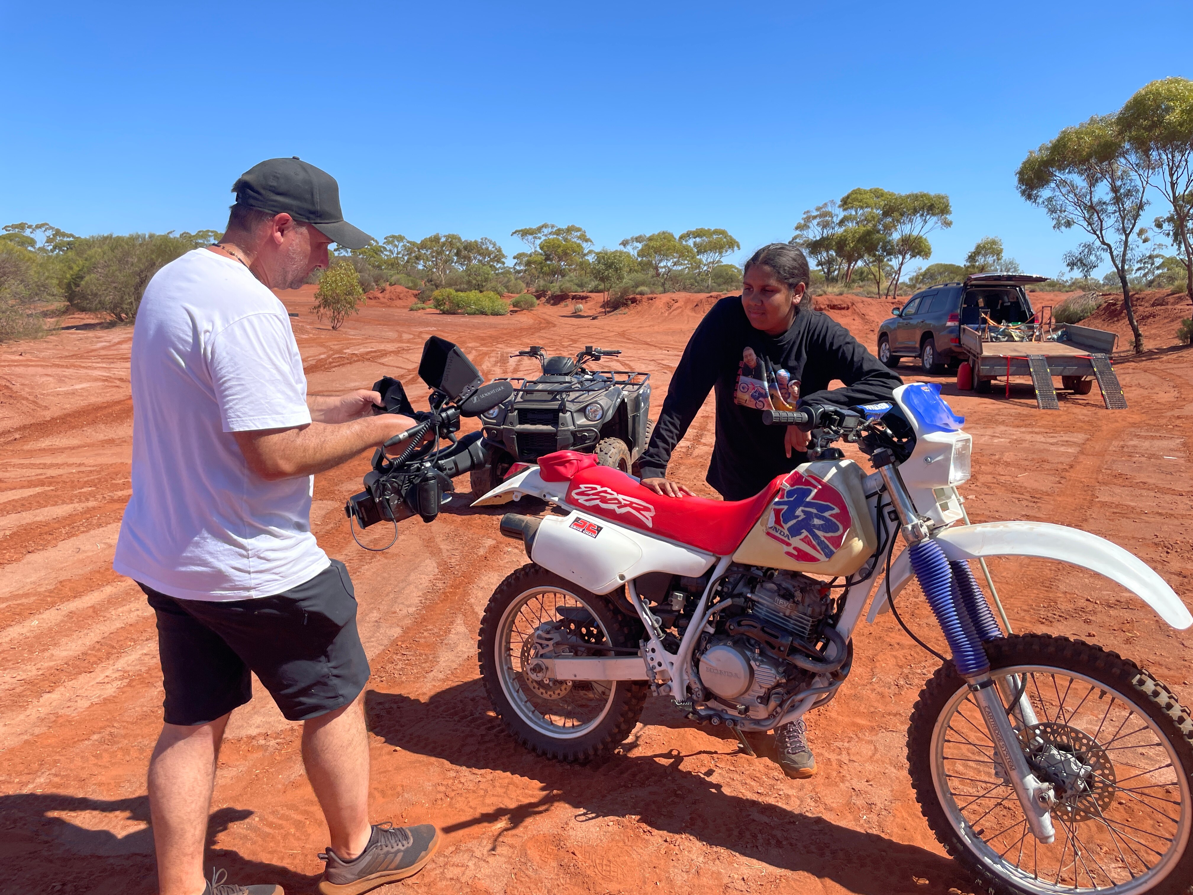 Uma jovem está atrás de uma bicicleta suja vermelha, azul e branca enquanto um homem segura uma câmera filmando-a.