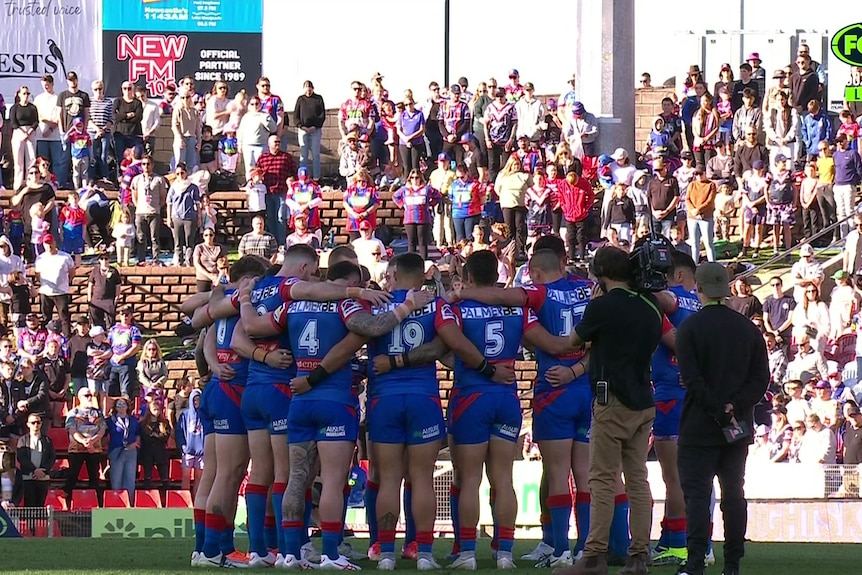 a group of nrl players grouped together in a circle arm in arm stand in front of a packed stadium