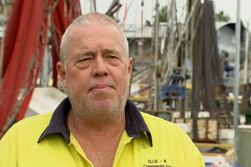 A man wearing a yellow collared shirt stands in front of fishing vessels.