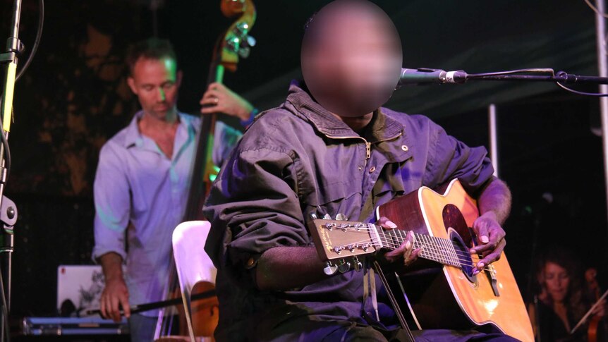 Dr G Yunupingu sits on stage performing with a guitar with another musician on double bass behind him.