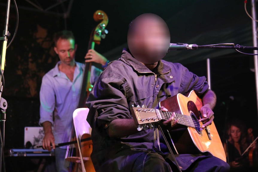 Dr G Yunupingu sits on stage performing with a guitar with another musician on double bass behind him.