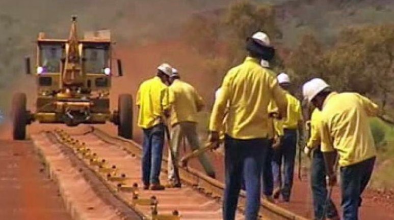 Construction workers wearing bright yellow shirts work on a rail line in the Pilbara, with a rail car in the background.