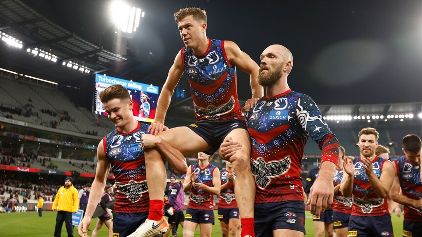 A Melbourne AFL player is carried off the ground on the shoulders of two teammates.