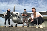 Two people standing and one squatting next to a small propeller plane