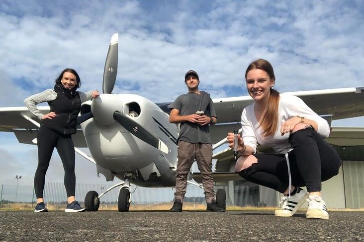 Two people standing and one squatting next to a small propeller plane