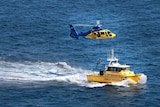 A helicopter winches someone down onto a rescue boat speeding along the ocean.
