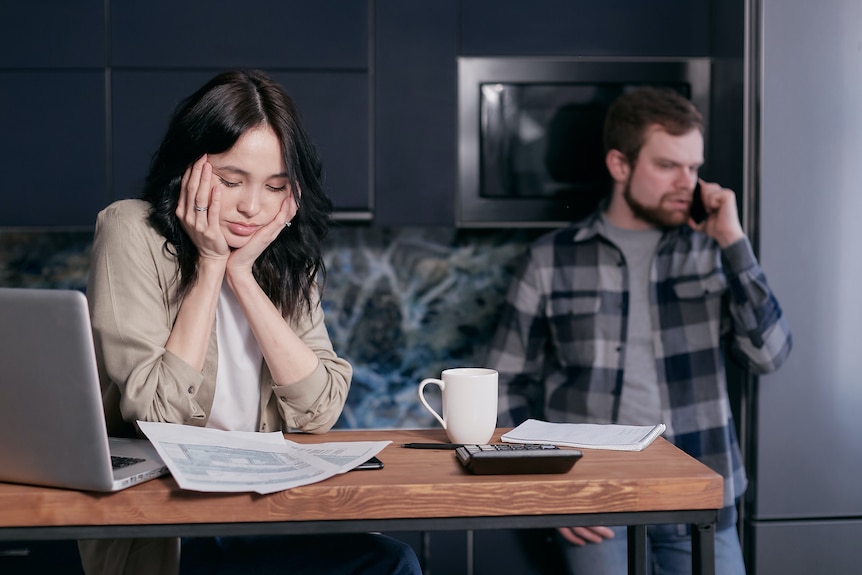 Woman sits at a desk looking at a calculator and bills, a man is standing behind her on the phone, both look stressed