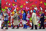 Women in gowns and masks line up with a backdrop of a painted red wall with hearts.