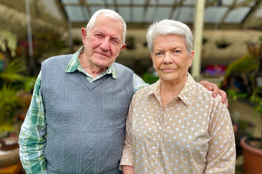 A couple with grey hair stands in a greenhouse, the man with his hand on the woman's shoulder.