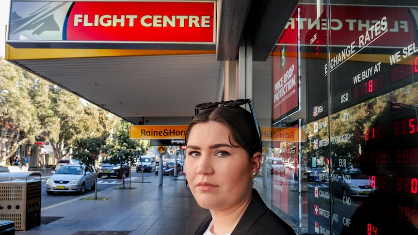 Olivia Little, wearing a black jacket and white shirt, stands on street in front of Flight Centre store.