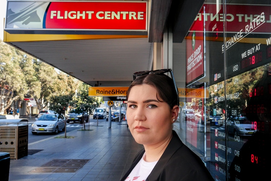 Olivia Little, wearing a black jacket and white shirt, stands on street in front of Flight Centre store.