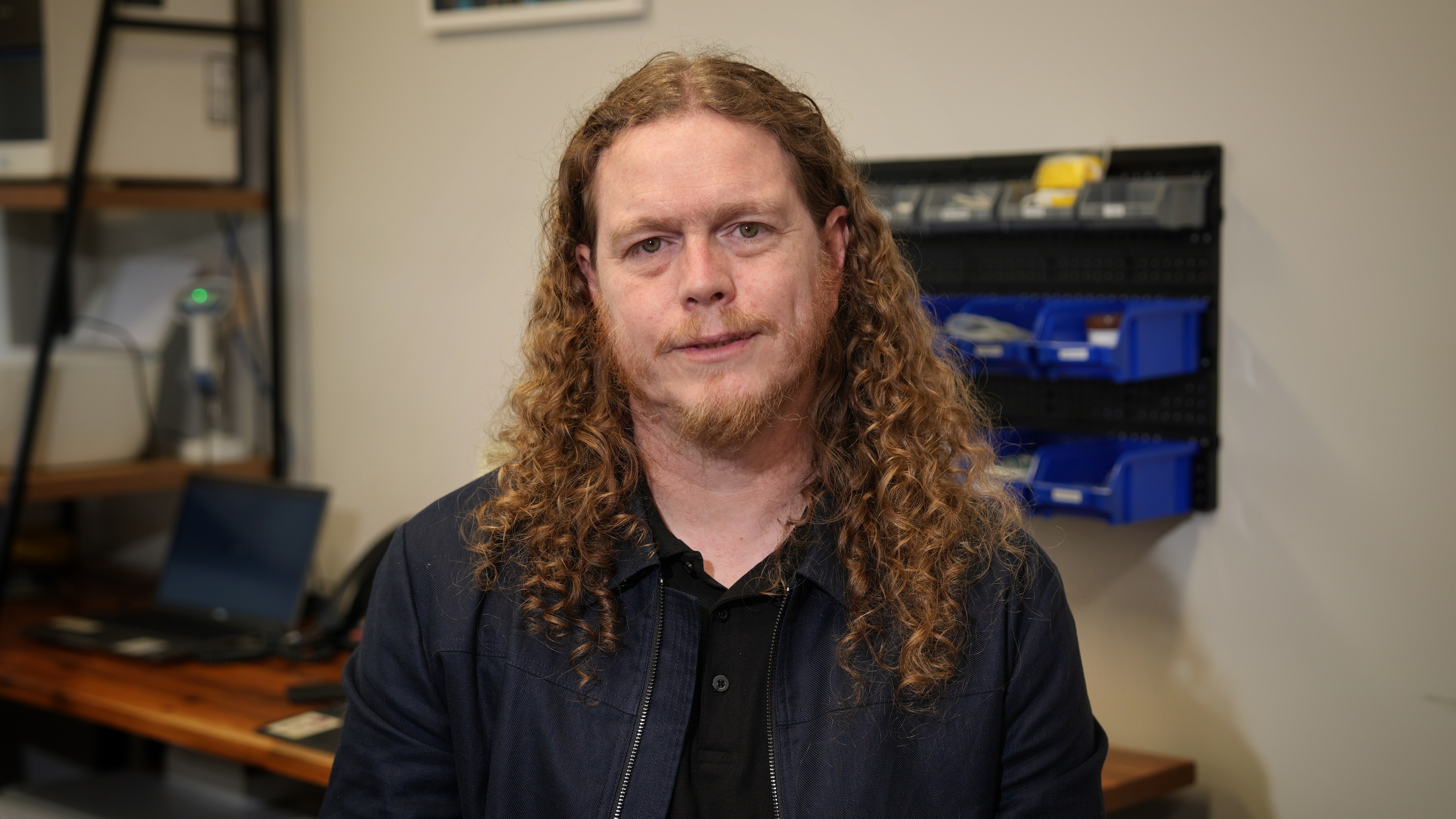 A man with long ginger hair sits in an office looking serious.