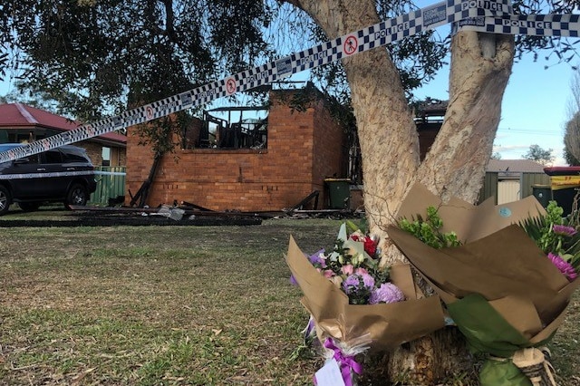 Bunches of flowers stacked up against a tree with police tape tied around it, which extends around a burnt-out house.