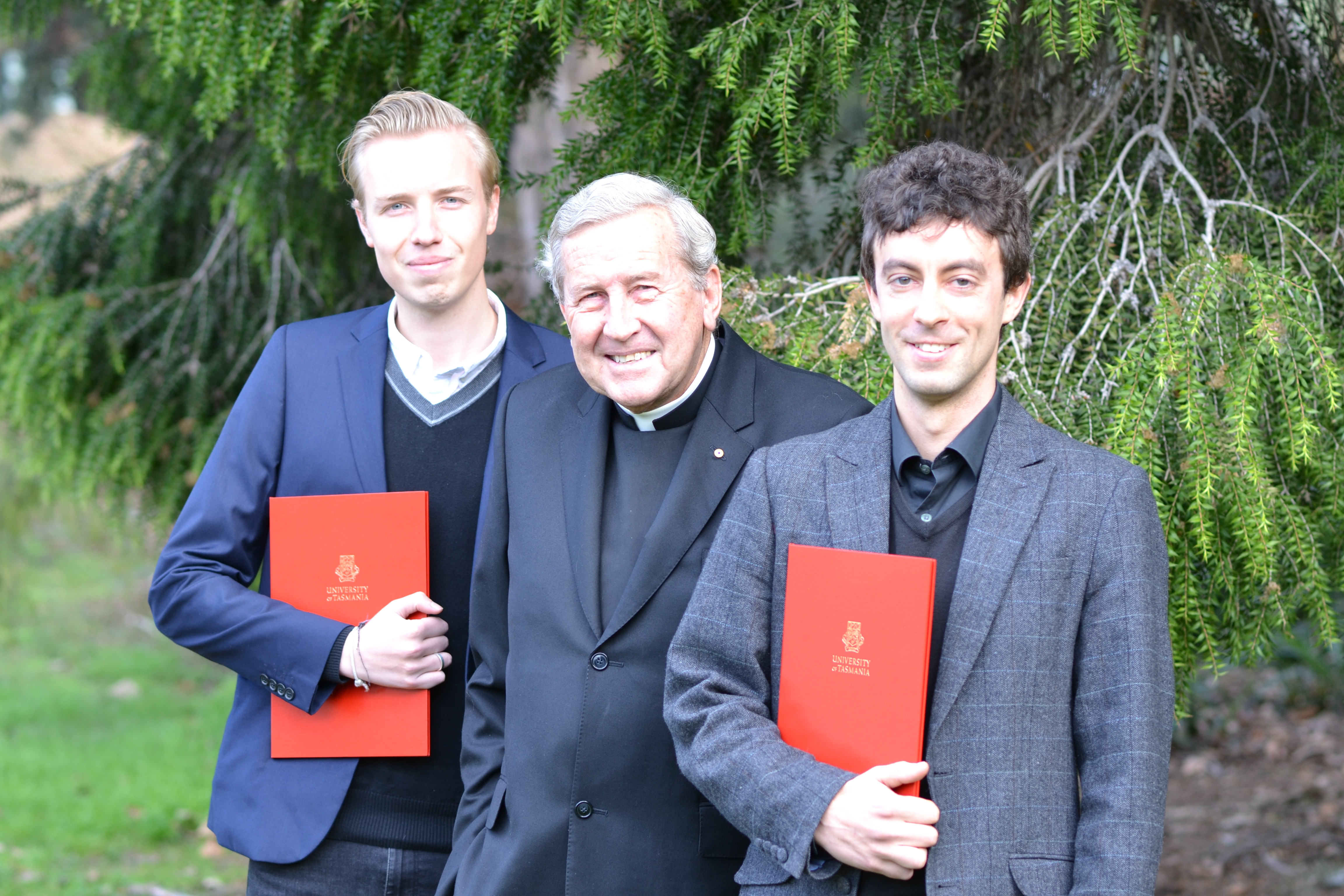 Michael Tate stands between two students who each hold a red folder with the University of Tasmania's logo on the front.