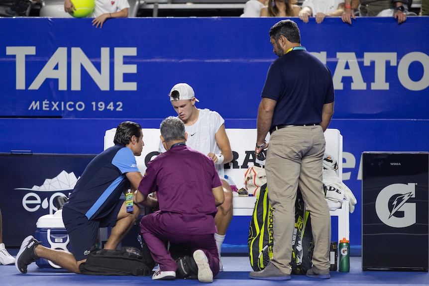 A tennis player sits on the sidelines, surrounded by medical staff, trainers and tournament officials during a medical break.