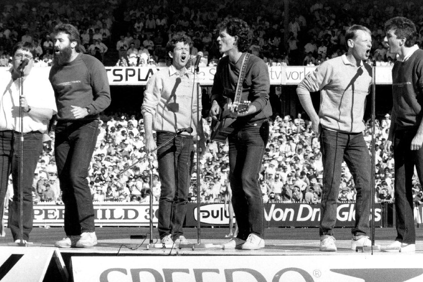Black and white photo of six men on stage singing into microphones on MCG with crowd in background.