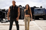 A muscly black man in black shirt and pants and a white woman in black crop and bleached jeans stand in front of large vehicles.