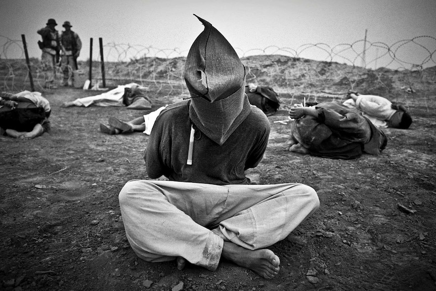 A Iraqi prisoner of war sits hooded surrounded by other POWs