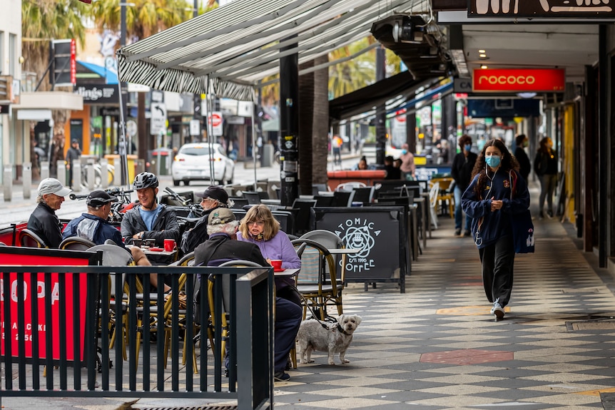 People enjoy a coffee outdoors as pedestrians walk past.