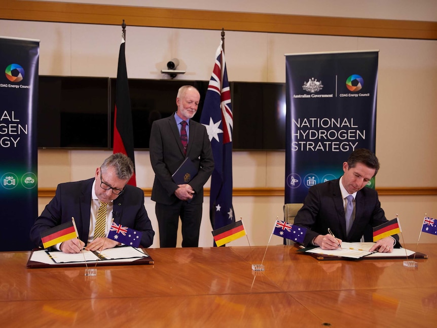 Two men sit at a large wooden table signing a document in front of little australian and german flags.