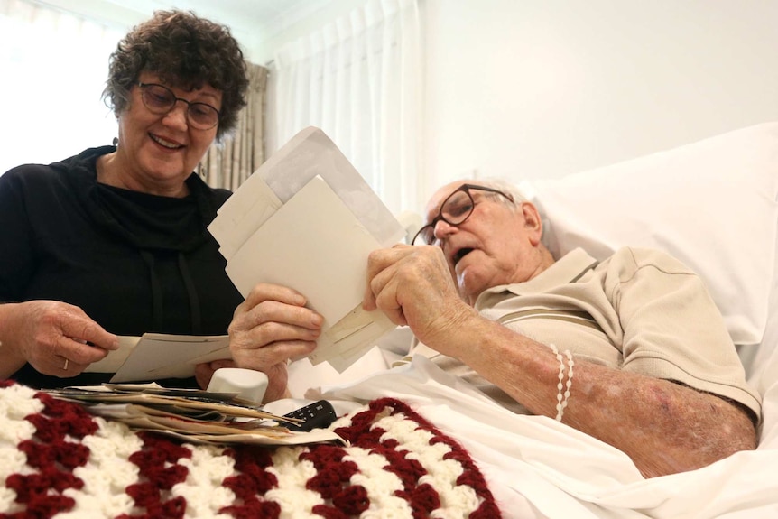 Woman with glasses sits next to an elderly man in a bed, both looking at photos.