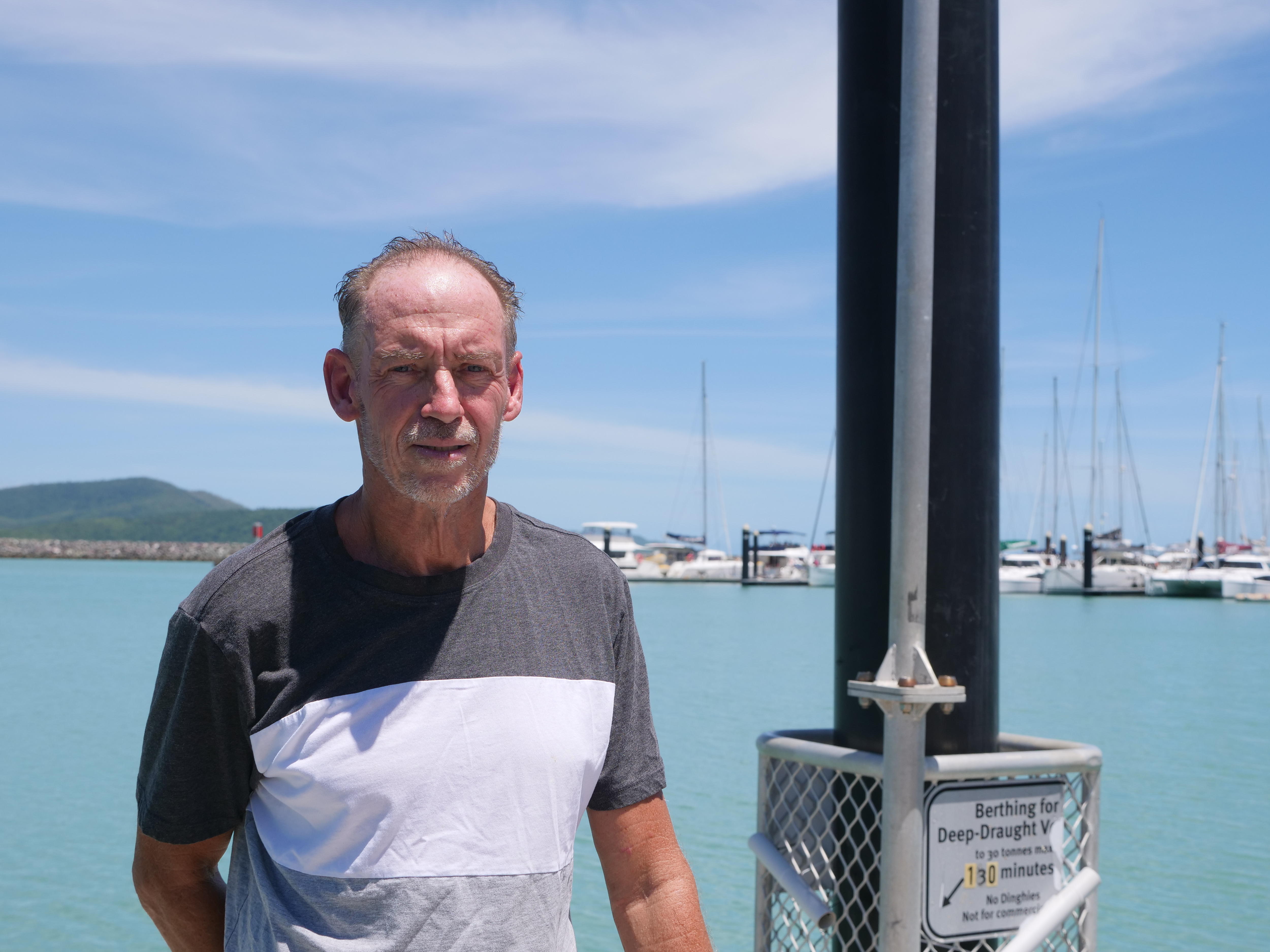 Un hombre de unos sesenta años con pelo corto se para frente a aguas poco profundas y barcos en un puerto deportivo. 