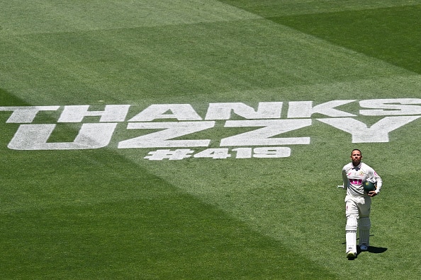 An aerial view of a cricket field showing painted words 'Thanks Uzzy #418. Player in cricket whites and helmet holds bat.