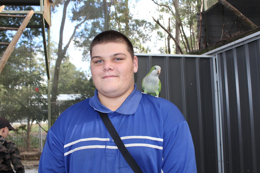 A teenage boy wearing a blue shirt with a greed bird perched on his left shoulder, standing in front of shed and trees