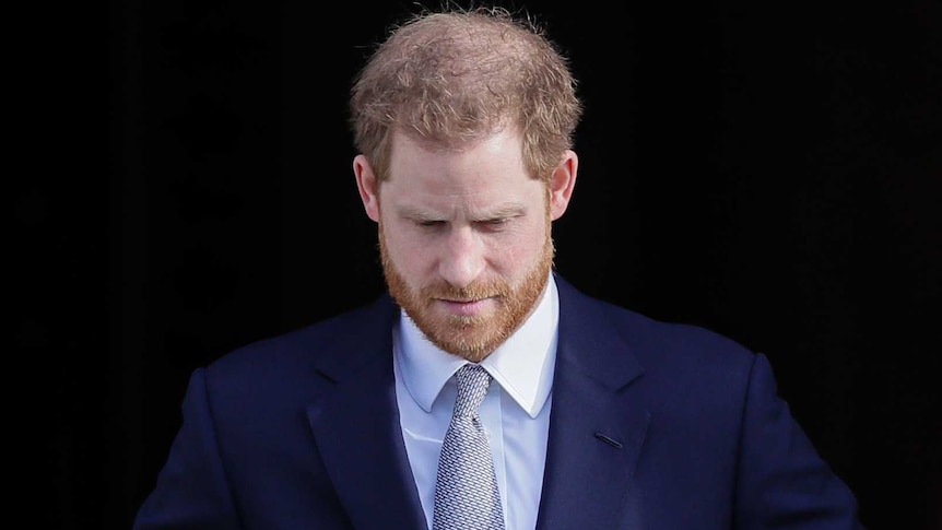 Prince Harry looks down as he fiddles with his jacket buttons while walking through a doorway.