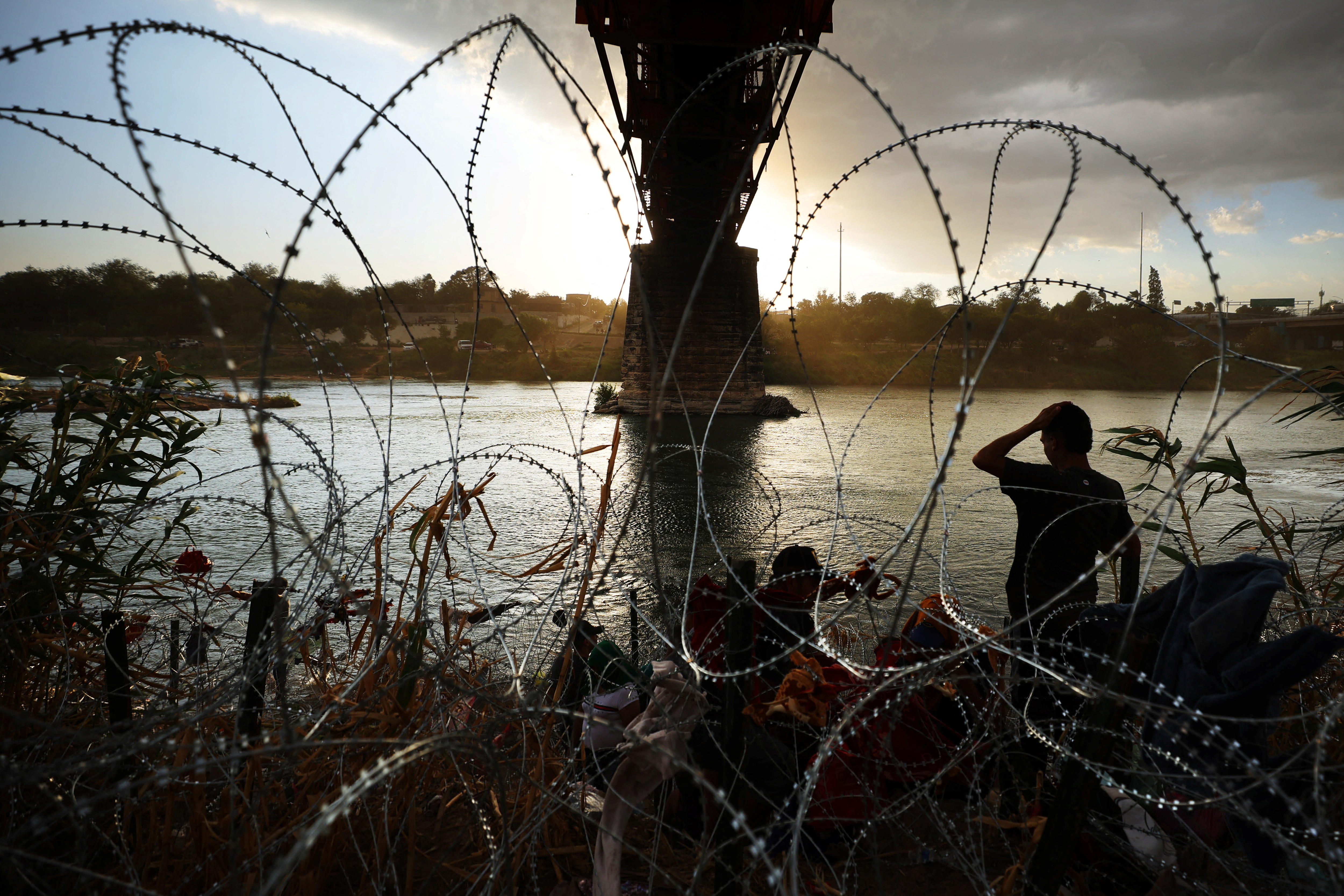 A silhouetted person stands behind loops of barbed wire on the banks of a river.