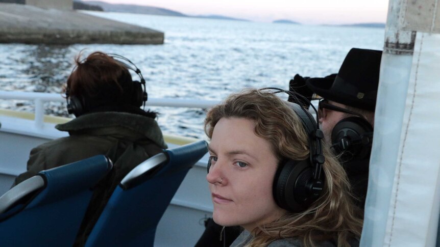 On at boat a young woman, wearing headphones. stares out onto the water, wearing headphones