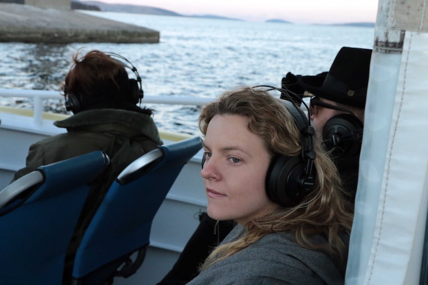 On at boat a young woman, wearing headphones. stares out onto the water, wearing headphones