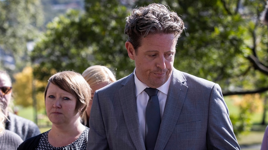 A man in a grey suit and his family stand in front of journalists.