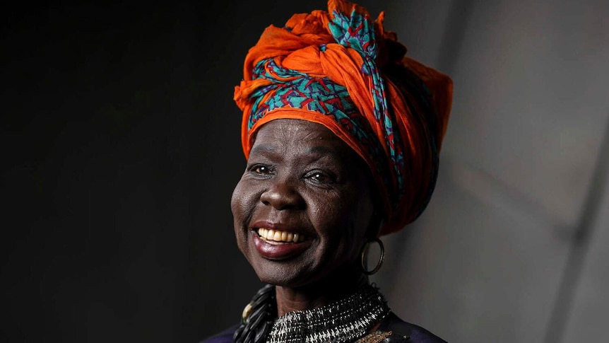 Ajak is smiling and looking away from the camera. She has an orange scarf in her hair, gold earrings and a large silver necklace