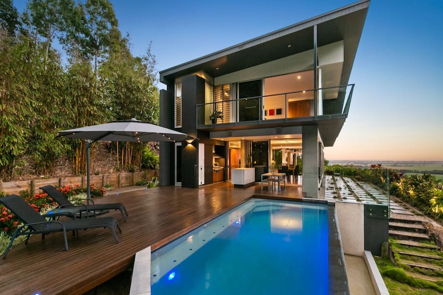 A two-storey hillside home with slanted roof overlooks the countryside out to the ocean. It has a narrow pool in the foreground.