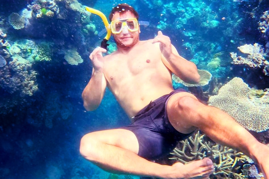 A young man, wearing blue boardshorts, goggles and a mask gives the thumbs up among coral.