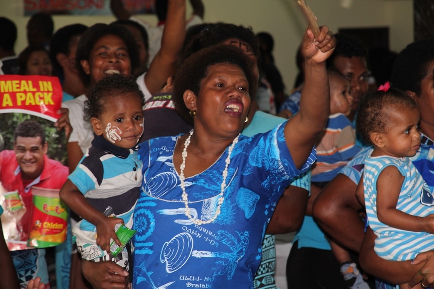 A mother cheers and holds a flag in the air while holding her toddler with one arm.