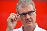 Michael Daley, in front of a red background, reaches for his glasses.