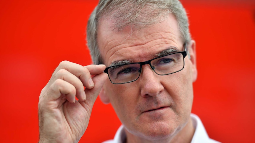 Michael Daley, in front of a red background, reaches for his glasses.