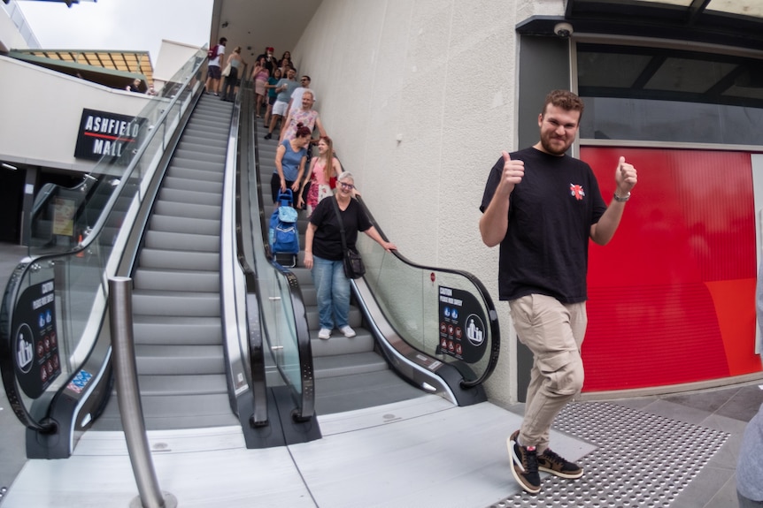 A young man steps off an escalator smiling, with two thumbs up. 