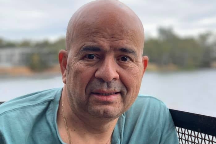 A portrait photo of a man in a blue shirt sitting on a balcony in front of a body of water.