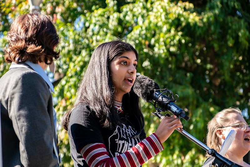 A girl with long dark hair, black t-shirt, stands at a microphone and speaks.