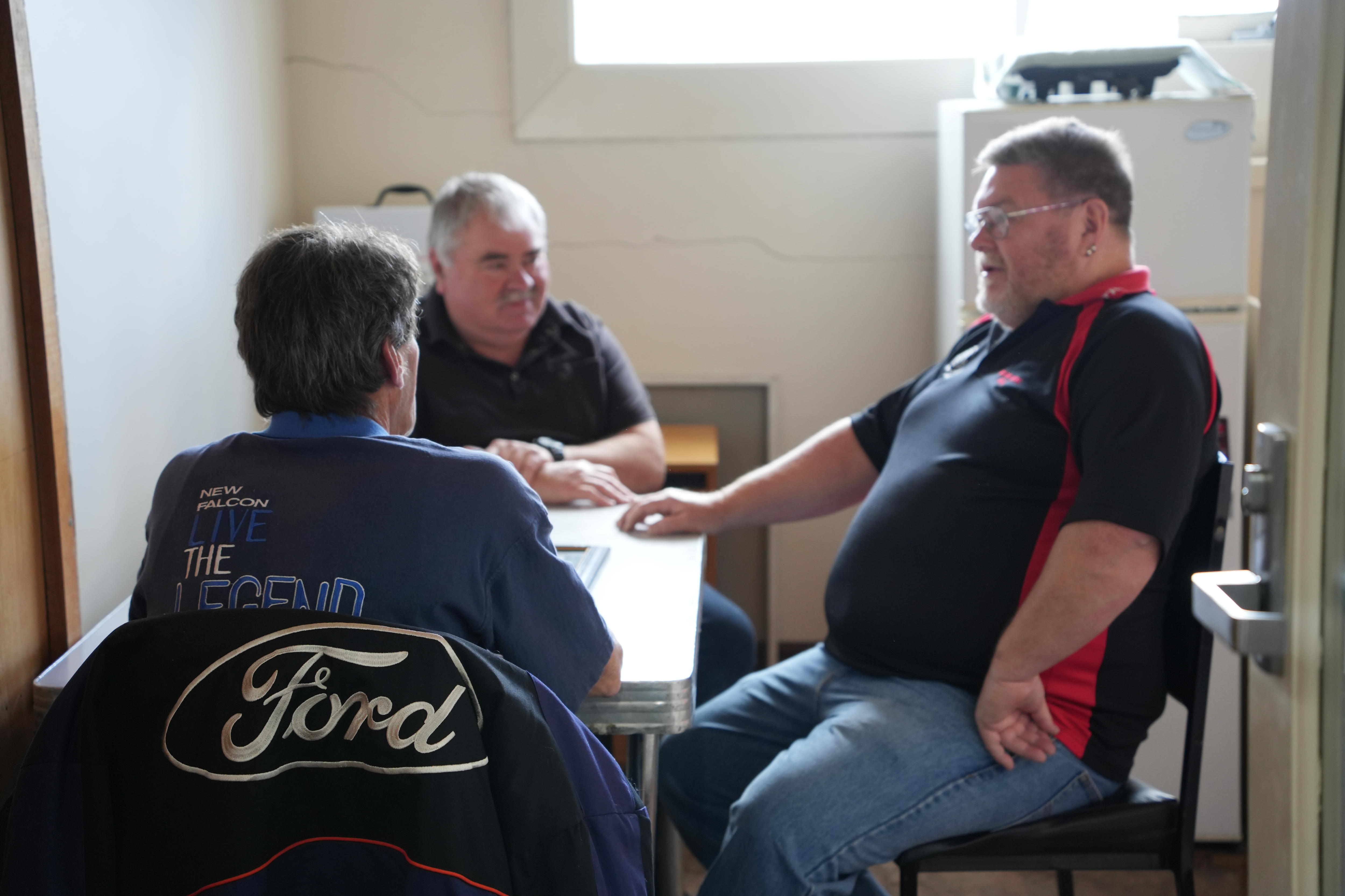 Three casually dressed middle-aged men sit at a table in a small factory team room.